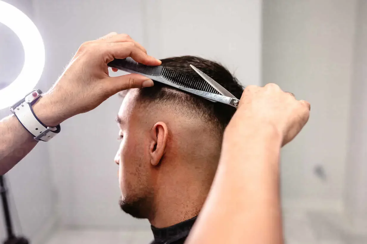 Close-up of a barber’s hands combing and cutting the hair on the back of a man’s head in a white-walled salon with circular light.