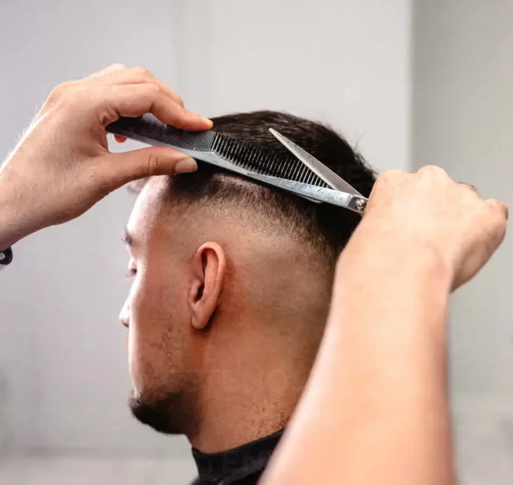 Close-up of a barber’s hands combing and cutting the hair on the back of a man’s head in a white-walled salon with circular light.