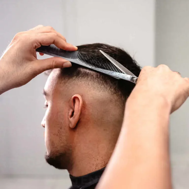 Close-up of a barber’s hands combing and cutting the hair on the back of a man’s head in a white-walled salon with circular light.
