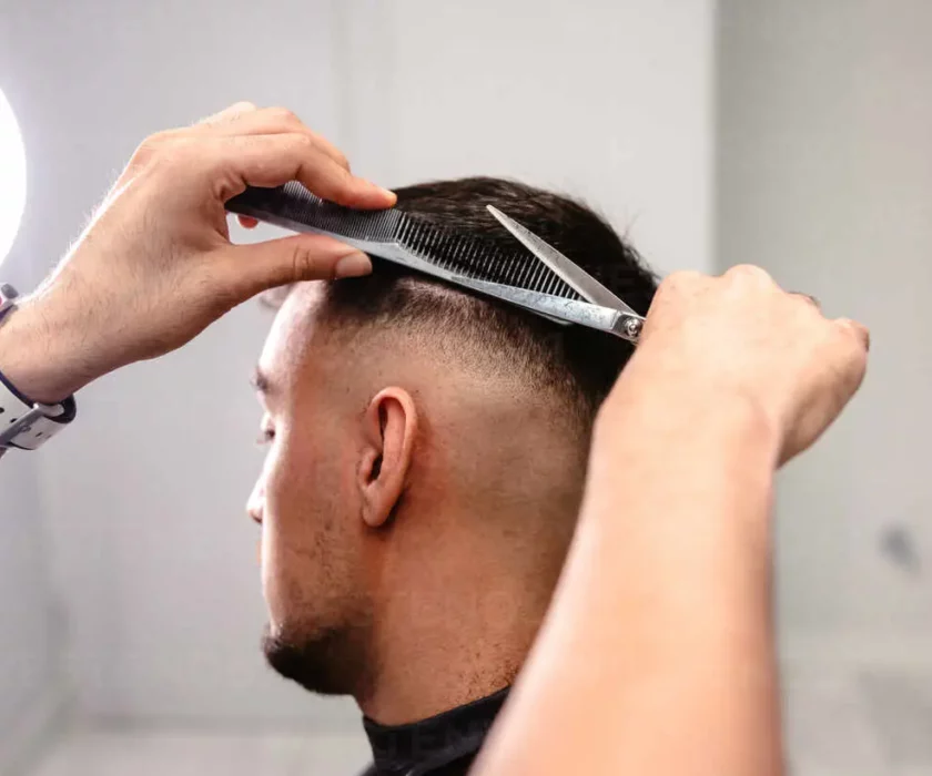 Close-up of a barber’s hands combing and cutting the hair on the back of a man’s head in a white-walled salon with circular light.